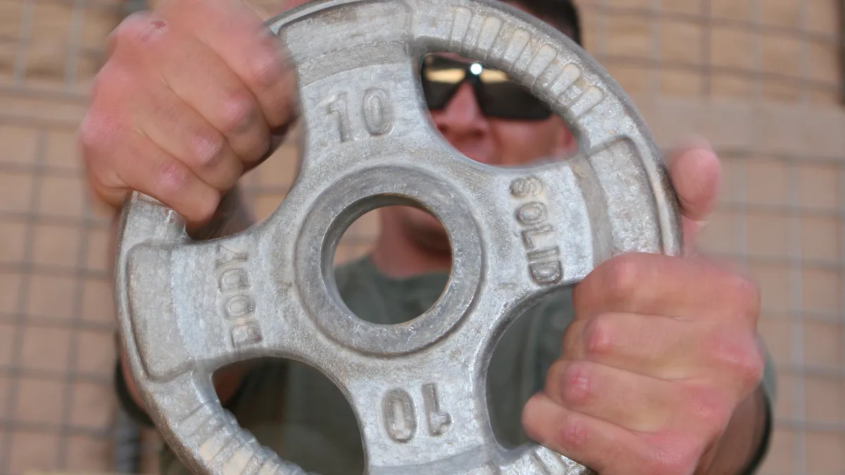 Two hands holding a 10-kilo weight plate in front of the face of an athlete wearing sunglasses